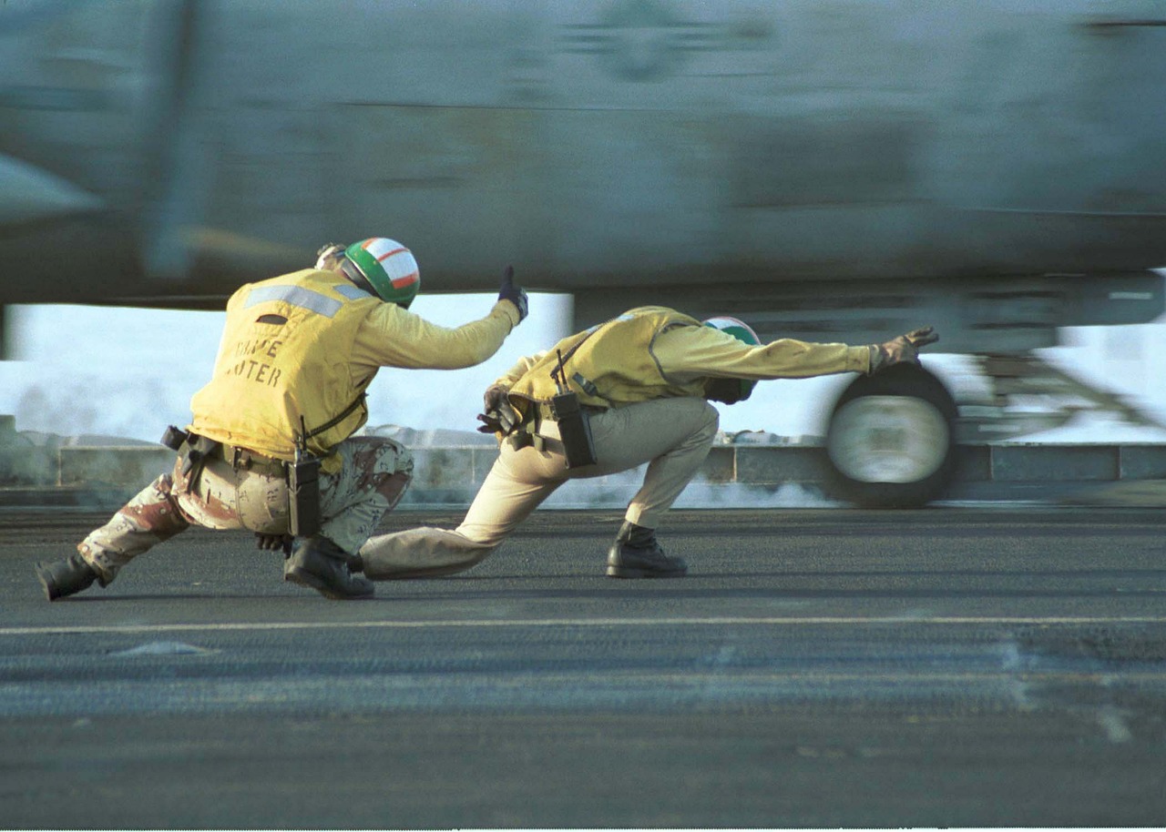 Two aircraft carrier crew members in yellow shirts and helmets crouch on the runway, guiding a jet during launch operations with hand signals.