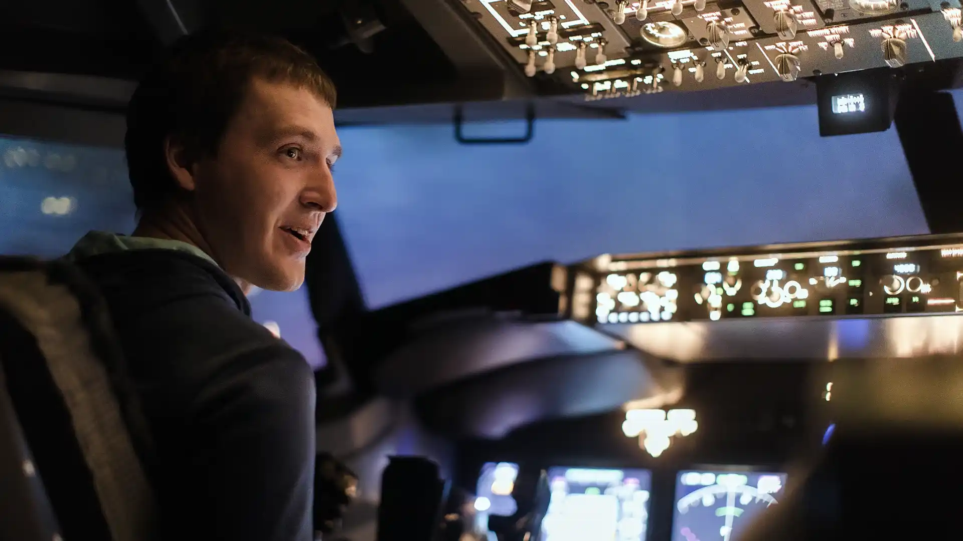 A man is seated in an aircraft cockpit with the dashboard illuminated in the background.