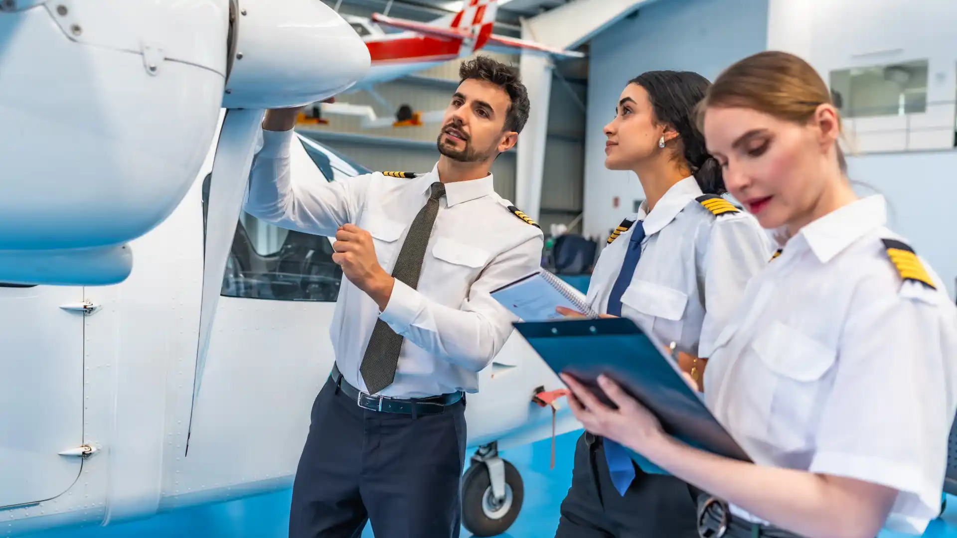 Three airline pilots in white uniforms are conducting a pre-flight inspection of a light aircraft’s engine and propeller.