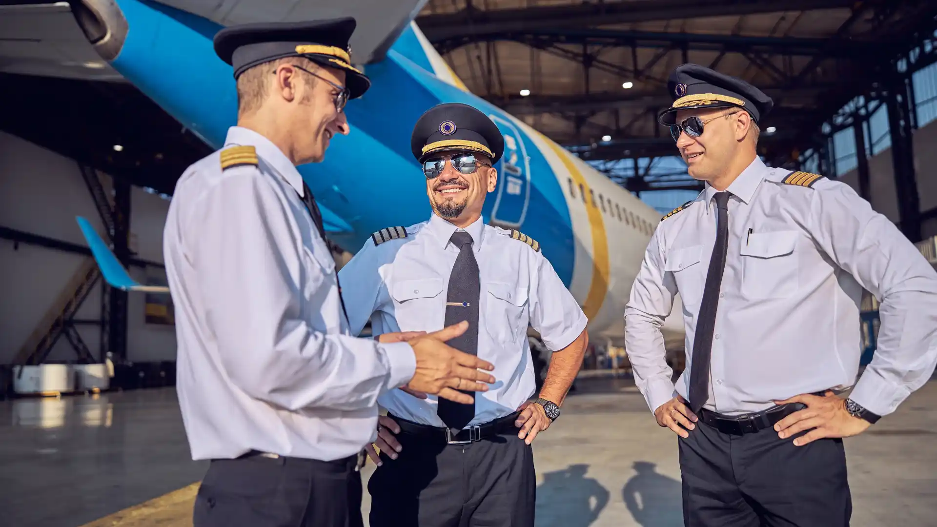 Three airline pilots in uniform shirts and caps talk in an aircraft hangar with an airplane in the background.