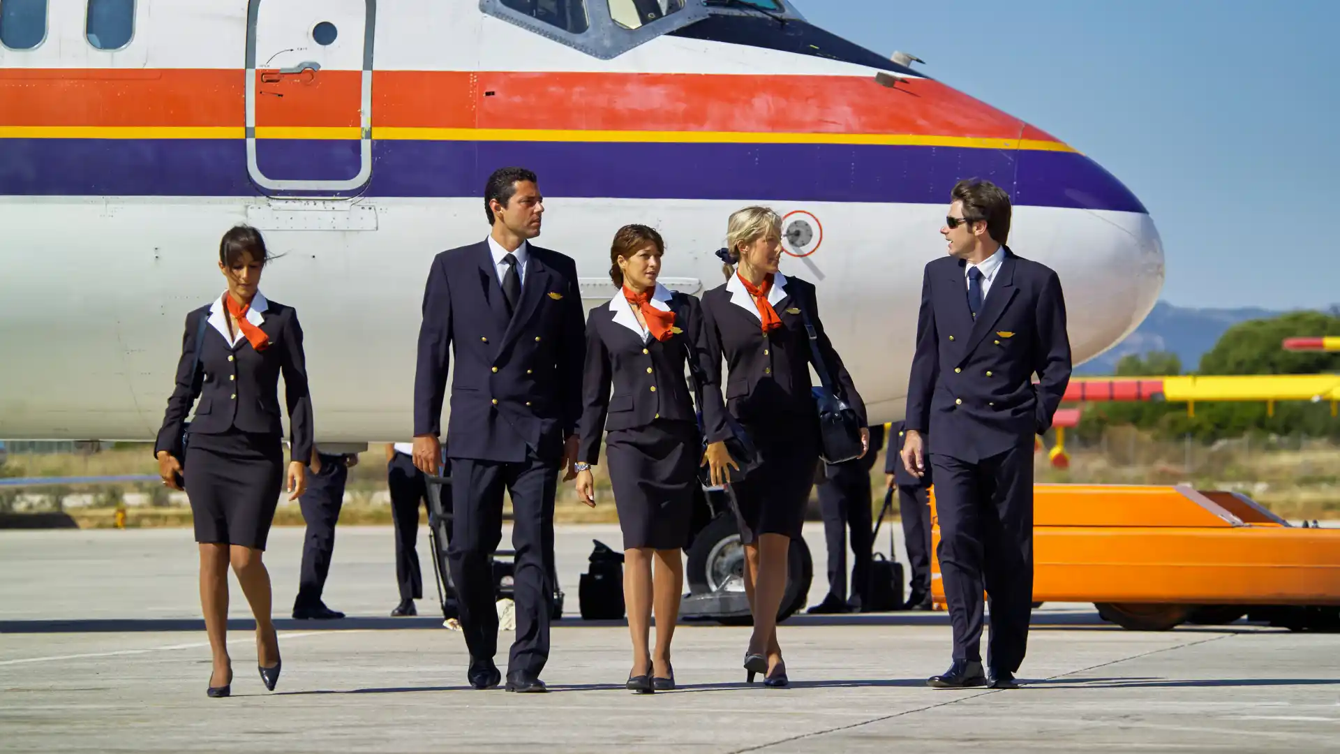 A flight crew, including pilots and cabin crew, walking together on the tarmac in uniform beside a passenger aircraft.