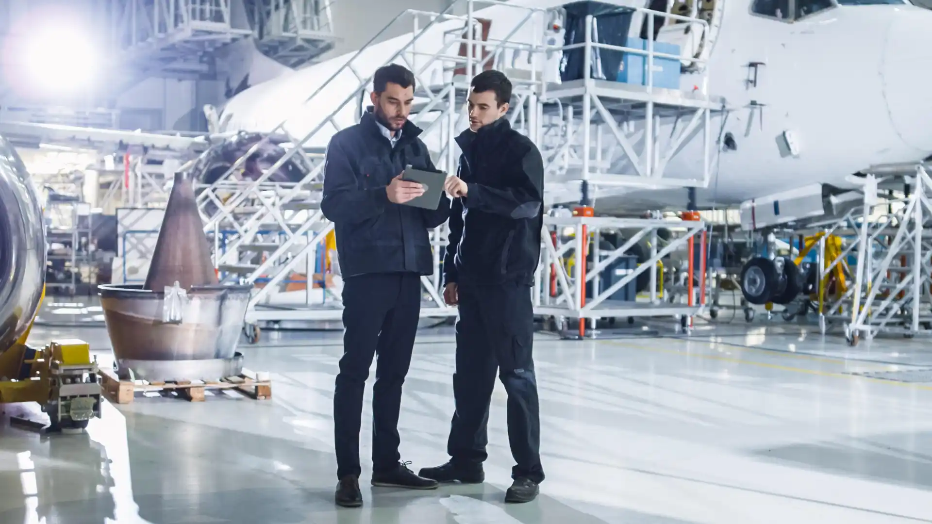 Two aircraft maintenance engineers in a hangar discussing information on a tablet, with an aircraft undergoing detailed inspection and servicing in the background.