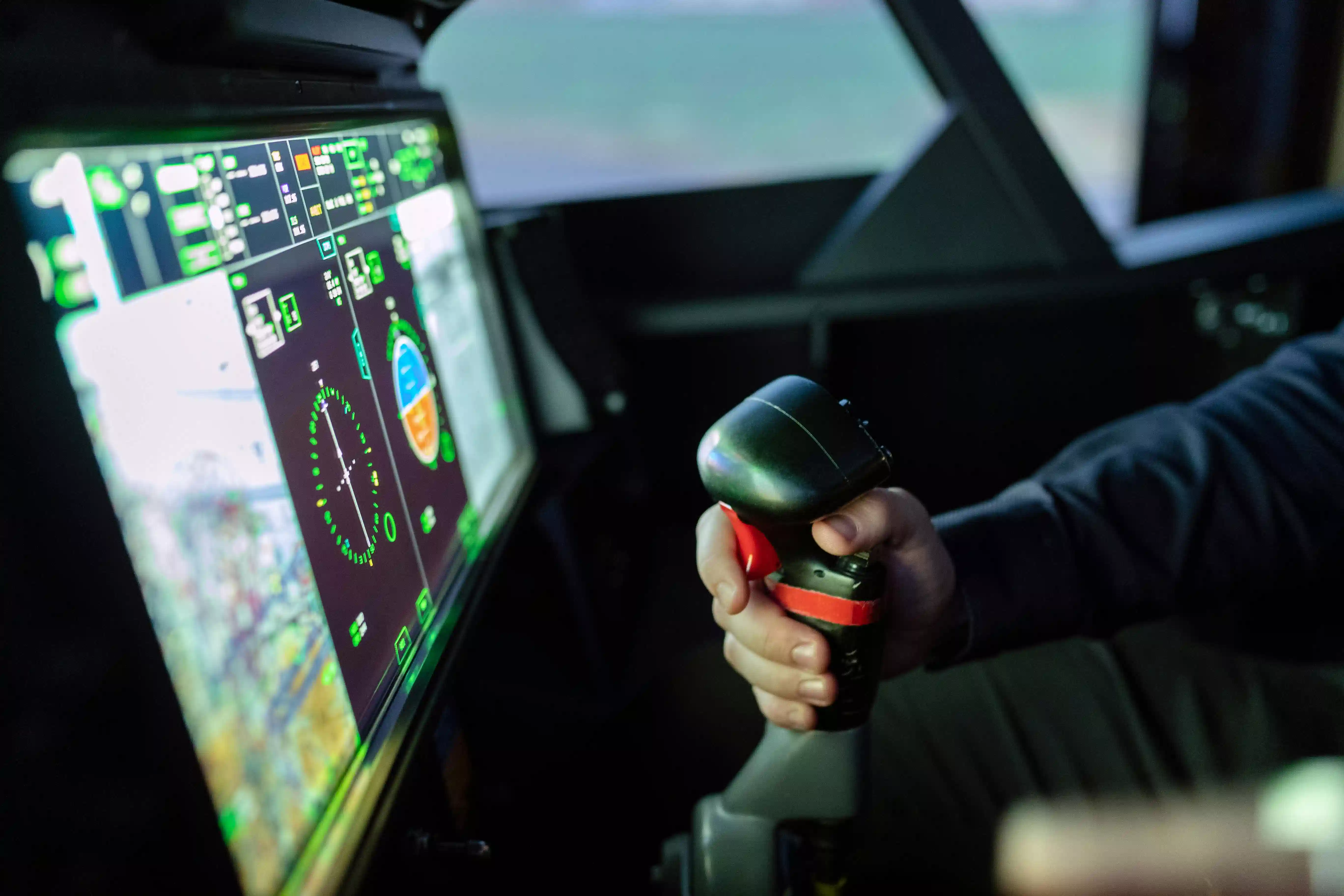 A pilot operates a joystick in a darkened cockpit with advanced digital flight displays.