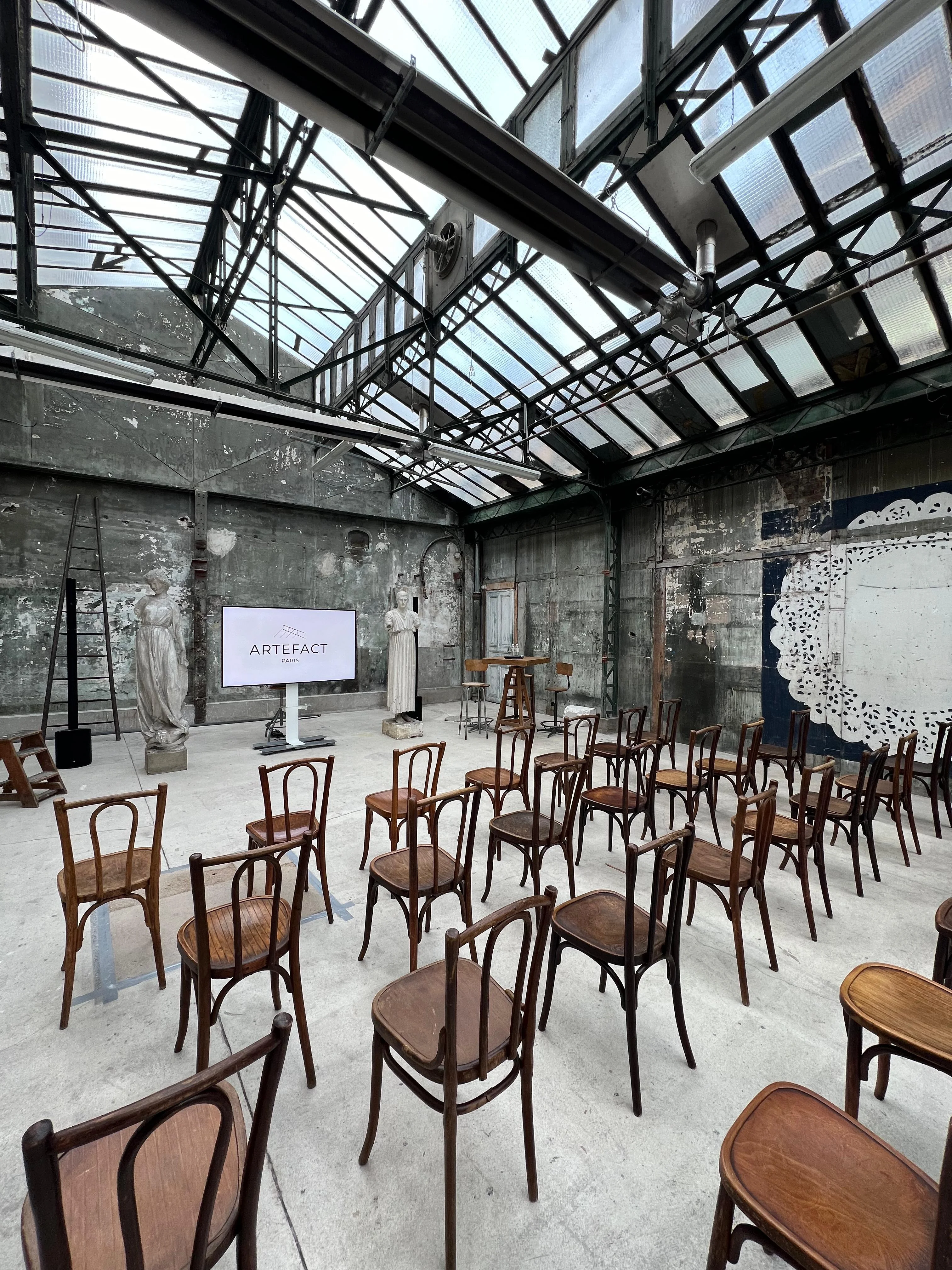 Salle polyvalente aménagée avec des tables et chaises blanches, éclairée par des suspensions industrielles, prête à accueillir des réunions ou des ateliers dans un cadre atypique avec un sol en bois ancien