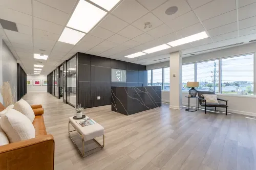 Calibrex's office reception with a black marble desk, dark wood accent wall, and light wood flooring. A caramel leather couch with white pillows sits on the left, and large windows on the right brighten a cozy seating area with a chair, table, and lamp.