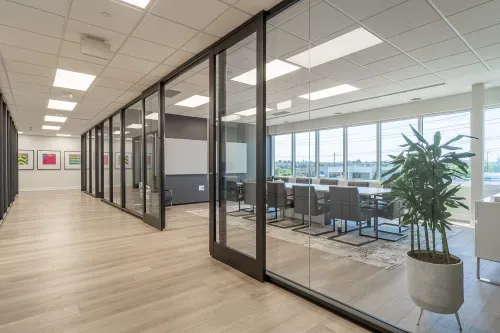 A long hallway with light wood floors that features panes of glass that section off office spaces. Large bright windows light a large modern boardroom with contemporary grey chairs surrounding a large table. A potted plants and art decorate the area.