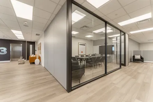 A modern office space featuring a glass-enclosed conference room with a large table and black chairs. The area has light wood flooring, with a cozy seating area to the left, including a mustard-colored couch and decorative accents. An elevator with a prominent floor number is visible in the background.