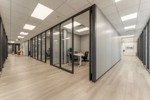 A modern office space featuring a glass-enclosed conference room with a small table and black chairs. The area has light wood flooring, with a cozy seating area to the left, including a mustard-colored couch and decorative accents.