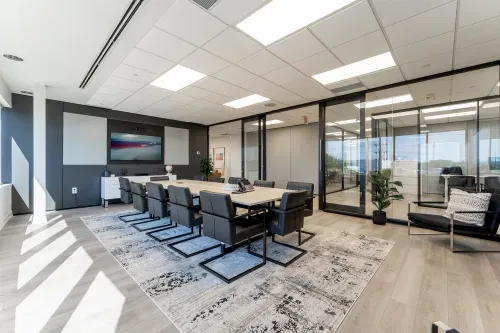 A large bright boardroom with light wood flooring, a wall with grey panelling and a wall of glass with large glass doors. In the center of the room is a large boardroom table surrounded by 12 modern leather square office chairs
