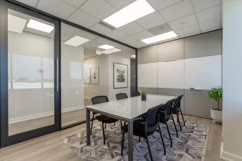 A modern office room with a glass table surrounded by 6 black office chairs. A grey and white panelled whiteboard wall is featured on the right wall and a glass wall with a door is on the left wall. The floors are light wood with a geometric carpet on top and plants on the able and in the corner of the room.