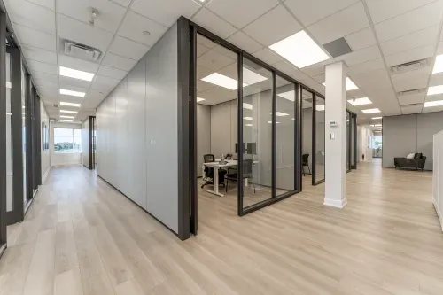 A large office space with three small offices in the center of the space and doors on the outside walls of the space. The floors are light wood, the walls a mix of grey panelling and glass.