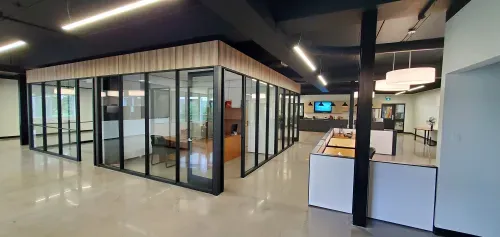 A large open office space with a pod of offices in the middle of the room seperated by glass walls. The floors are shiny grey concrete, the ceilings are black with modern lighting. To the left of the room is a open area of standing desks and at the back is a modern kitchen.