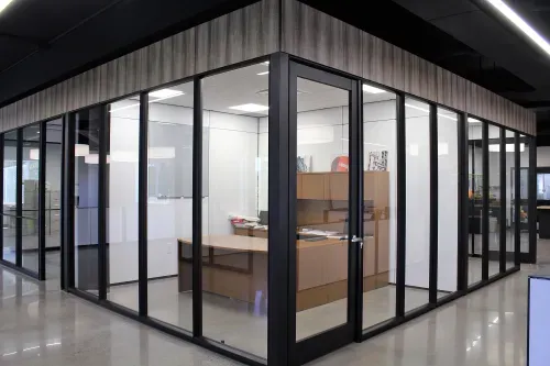 A corner office with glass walls and a wooden accent border along the top of the walls. Inside the office is a large wooden desk.