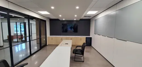 A large room with a whiteboard wall on the right, a black accent wall with a tv with a wooden cupboard under neither at the back and the left is a large glass wall with a door in it. Fold out tables and two office chairs are in the middle of the room.