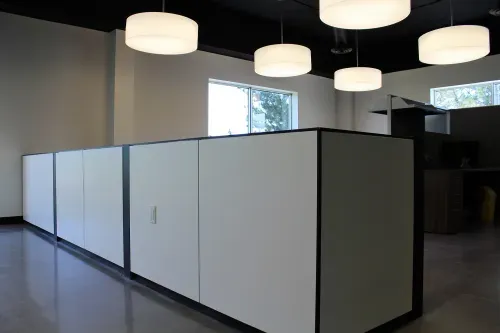 A large room divided why a white panelled half wall. Cylinder shaped white pendant lights hang from the ceiling.