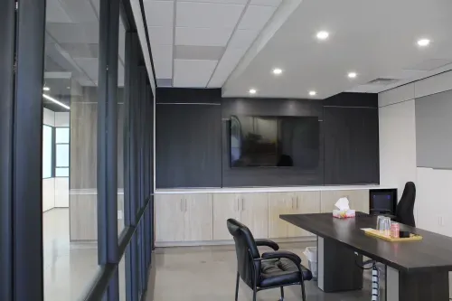 A small office with a single desk in the middle with chairs on either side. The back wall has black wood panelling and a mounted tv with cabinets under the tv.