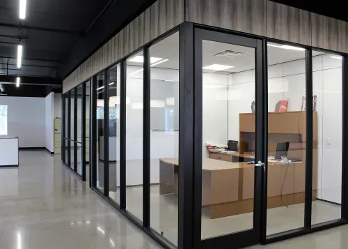 A corner office with glass walls and a wooden accent border along the top of the walls. Inside the office is a large wooden desk.