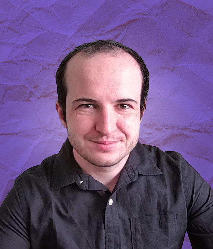 Photo of Quentin, a white man in his 20s with balding brown hair and stubble. They're wearing a black button-down shirt on a background of purple wrinkled paper. He's smiling.