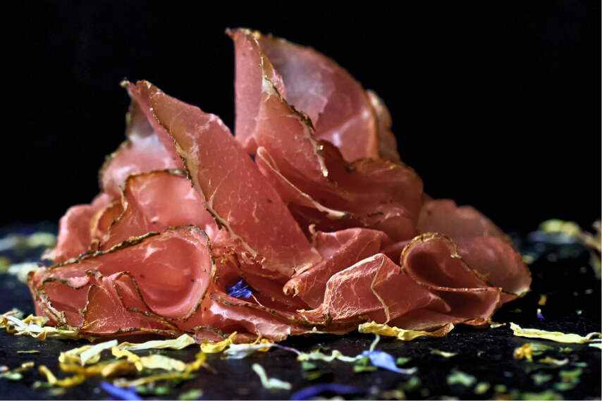 Fein geschnittene, luftgetrocknete Schinkenstücke auf schwarzem Untergrund mit verstreuten Blütenblättern.