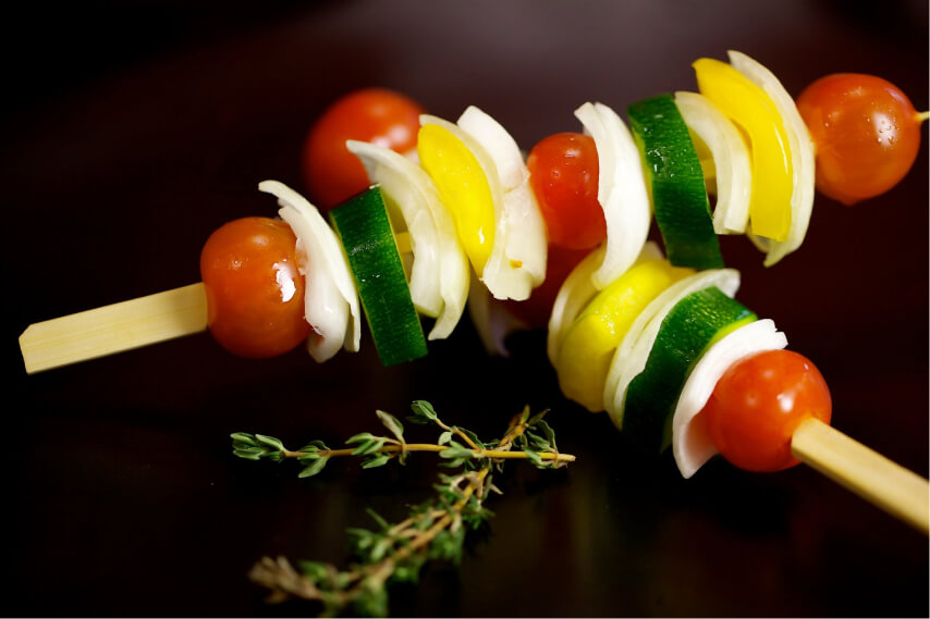 Zwei Gemüsespieße mit Kirschtomaten, Zucchini, gelber Paprika und Zwiebeln auf dunklem Hintergrund neben frischen Thymianzweigen.