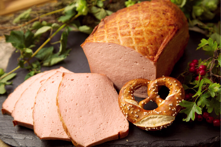 Frisch geschnittene Scheiben Fleischkäse neben einer Brezel auf einem schwarzen Steinbrett mit grünen Blättern und roten Beeren zur Dekoration.