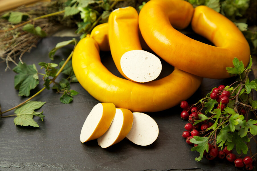 Mehrere Fleischwürste auf schwarzem Untergrund, davon eine angeschnitten mit sichtbarem weißem Inneren, dekoriert mit roten Beeren und grünen Kräutern.