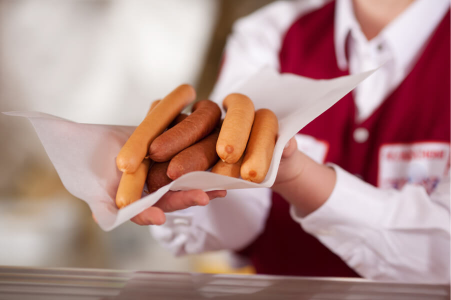 Metzgereifachverkäuferin reicht mehrere Würste auf einer Papierfolie über die Theke.