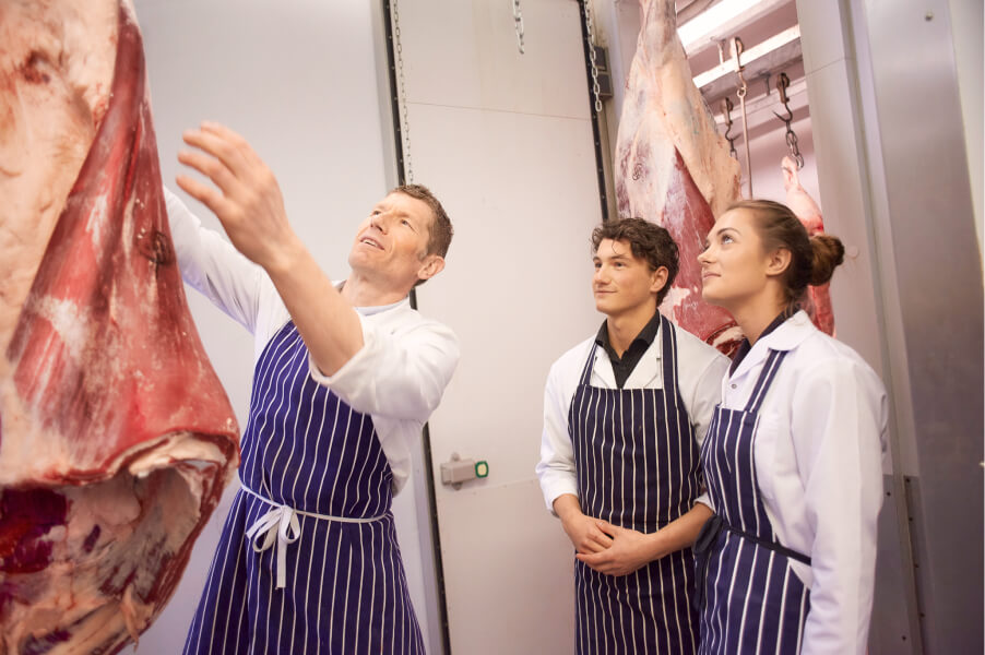 Drei Metzger in gestreiften Schürzen prüfen hängende Rinderhälften in einem Kühlraum.