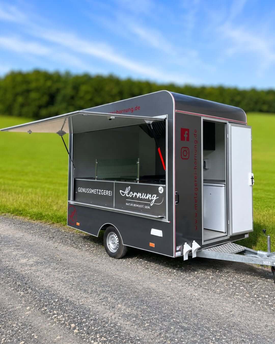 Grillwagen in grau mit Logo von Metzgerei Hornung auf Schotterweg. Im Hintergrund grüne Wiese und blauer Himmel