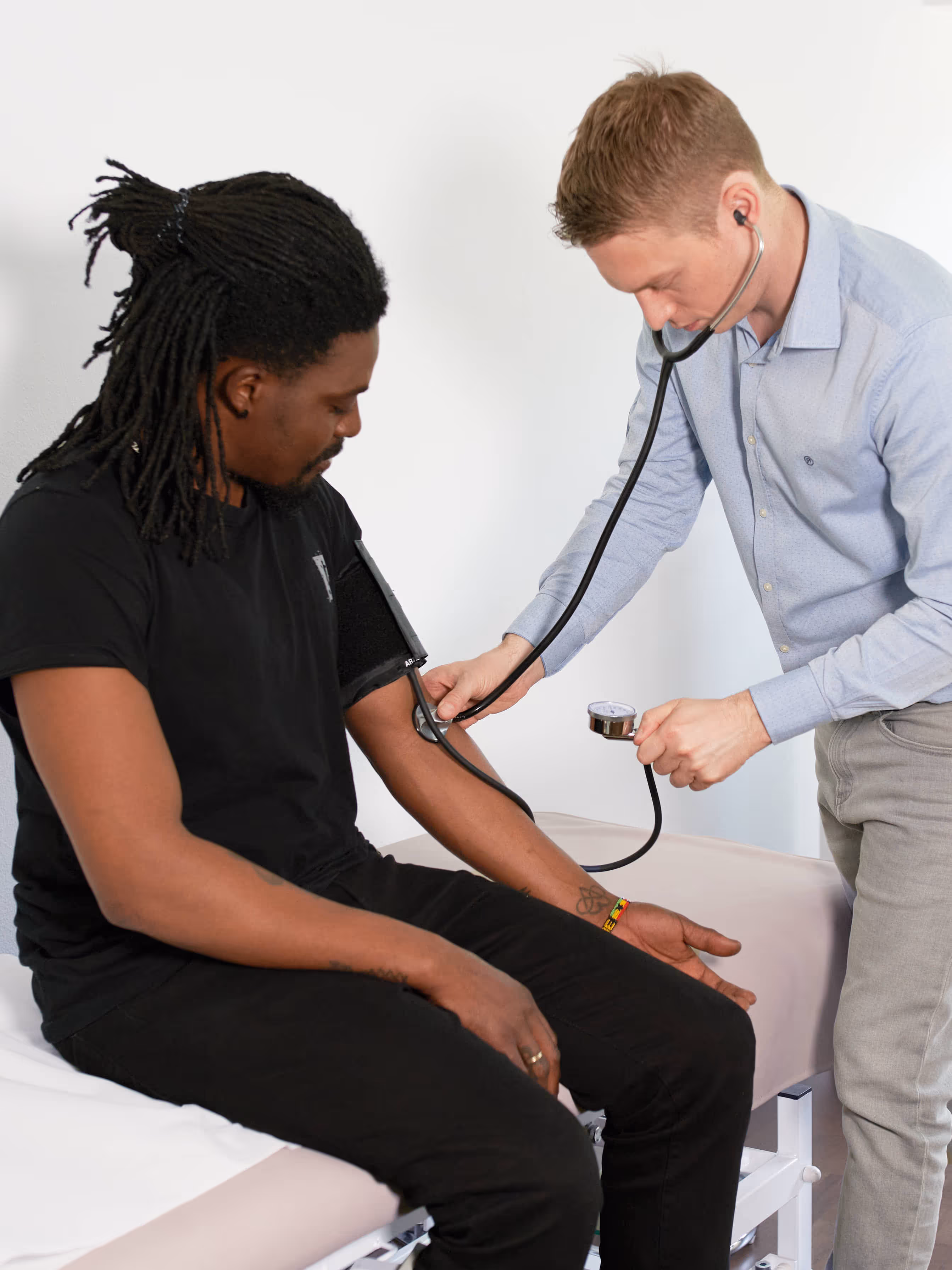 Ein junger Arzt mit Stethoskop hört einem sitzenden Patienten mit Dreads in schwarzem T-Shirt auf dem Herzen in einem hellen Behandlungsraum einer Praxis.