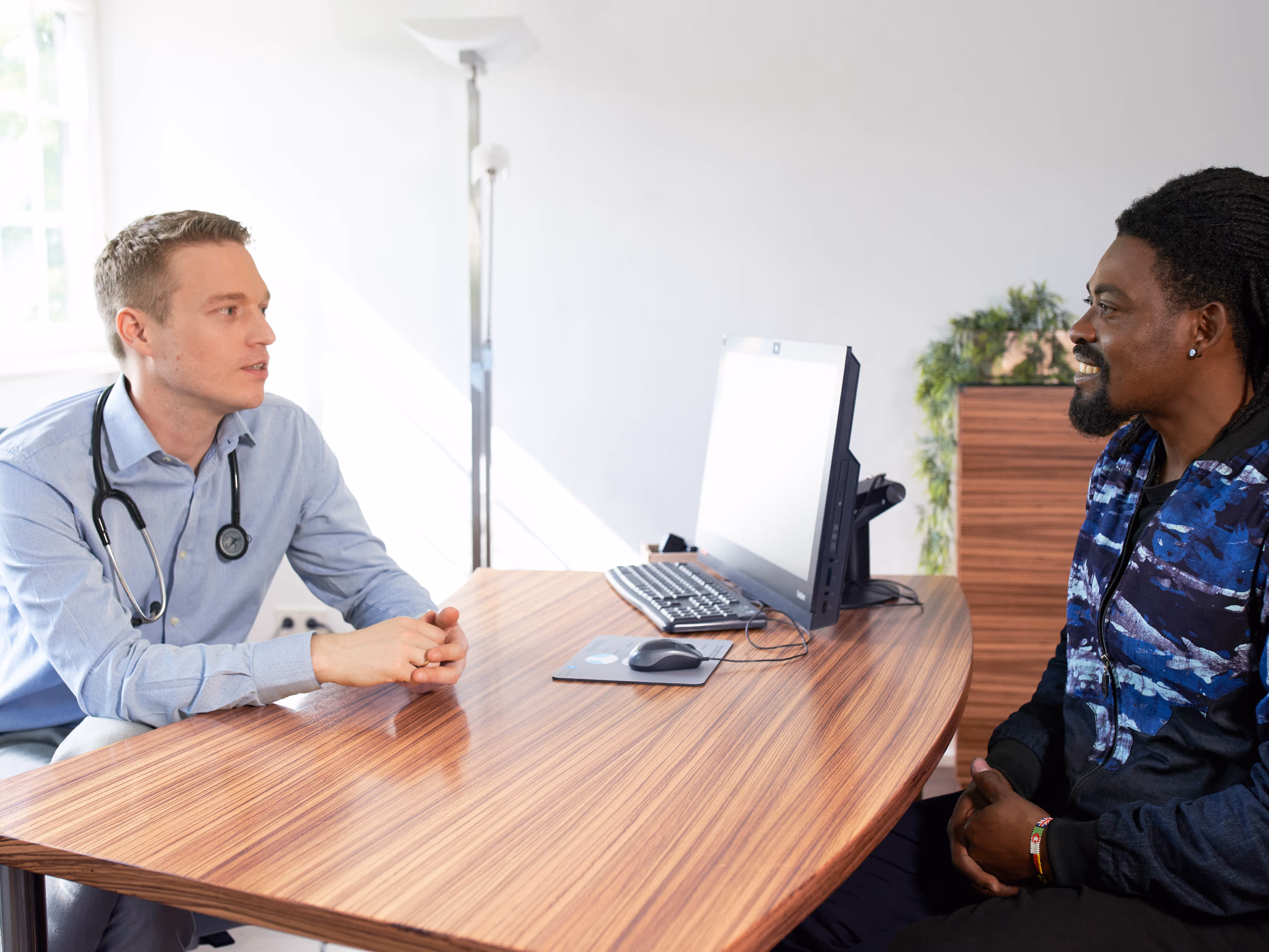 Arztgespräch: Weißer Arzt mit Stethoskop berät afroamerikanischen Patienten in blauem Jackett, entspannte Atmosphäre mit Pflanzen.