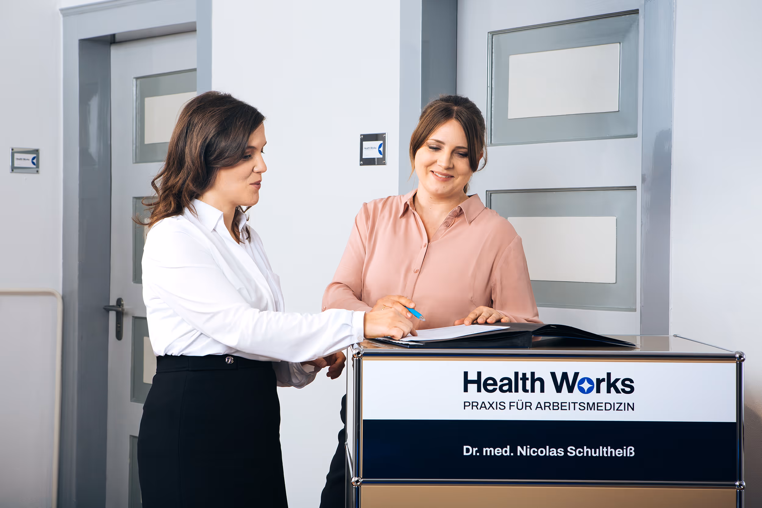 Zwei Frauen an der Rezeption von Health Works: Beratung in moderner Arbeitsmedizin-Praxis Dr. Nicolas Schultheiß.
