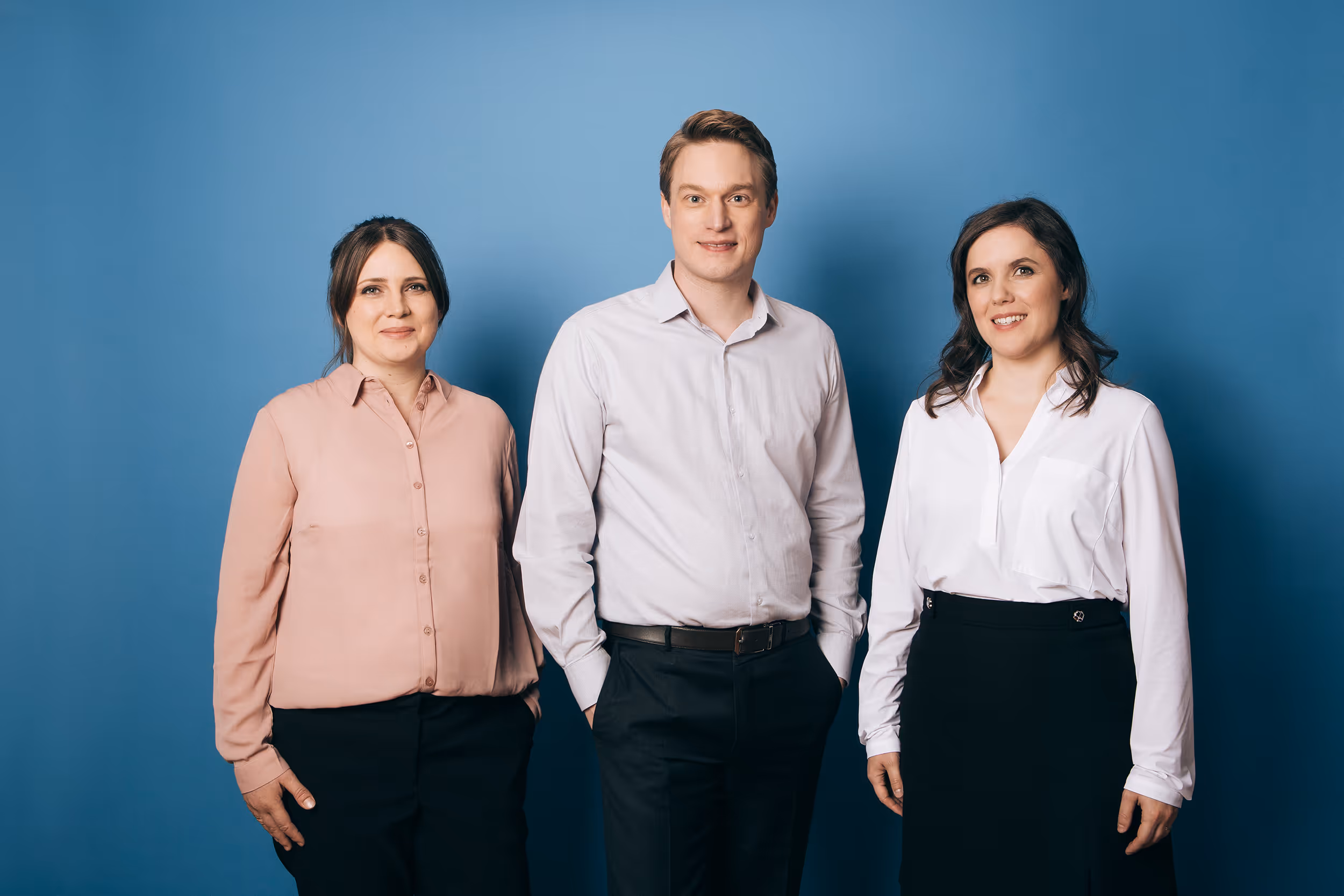 Drei Kollegen vor blauem Hintergrund: Frau in Rosa-Bluse, Mann in Hemd, Frau in Weißer Bluse – professionelles Teamfoto.