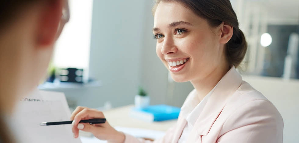 Jeune femme souriante en tenue professionnelle tenant un stylo lors d’une discussion au bureau.