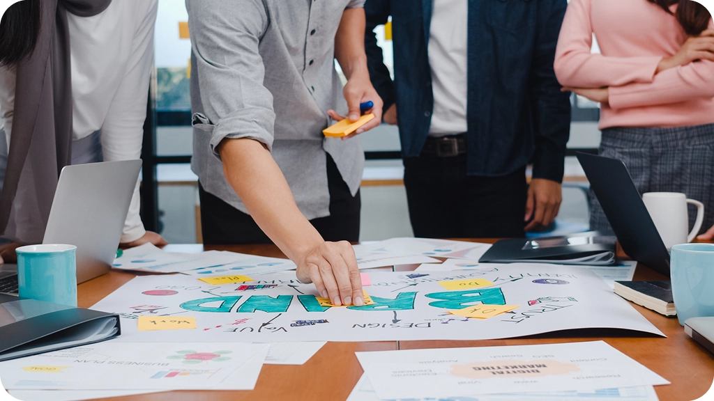 Un groupe de personnes autour d’une table de réunion travaillant sur une affiche de stratégie de marque (“Brand”). On voit des documents, des post-it colorés, des ordinateurs portables et des tasses. Une personne au centre colle un post-it sur l’affiche pendant que les autres observent.