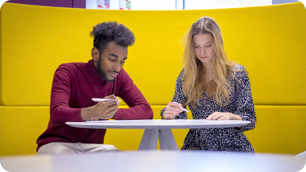 Deux étudiants concentrés travaillant ensemble à une table, assis devant une cloison jaune – BSB