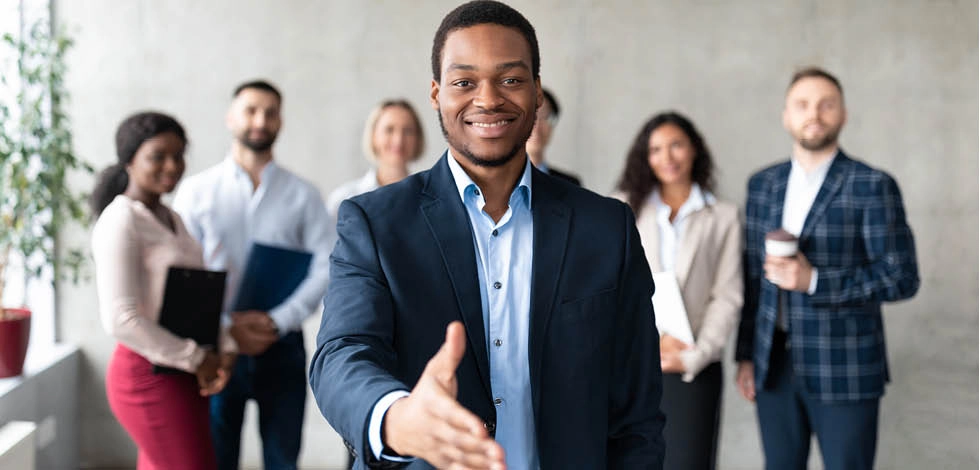 Un jeune professionnel en costume tend la main pour une poignée de main, entouré d’une équipe de collègues en arrière-plan, symbolisant le leadership, la gestion d’équipe et l’évolution de carrière vers le rôle de DRH – BSB