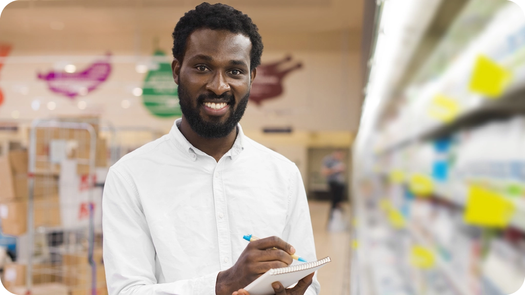 Jeune homme souriant en chemise blanche prenant des notes dans un supermarché, représentant les débouchés professionnels en gestion et commerce après une Licence AES – BSB