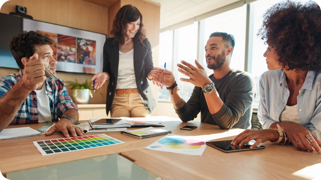 Un groupe de jeunes professionnels en pleine discussion autour d’une table de réunion, avec des échantillons de couleurs et des documents de travail, illustrant une séance de créativité en communication. - BSB