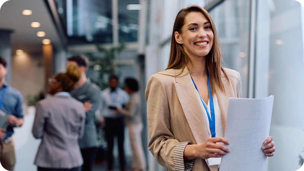 Une jeune femme souriante tenant des documents dans un couloir lumineux d’entreprise, portant un badge professionnel, tandis que d’autres collègues échangent en arrière-plan. - BSB