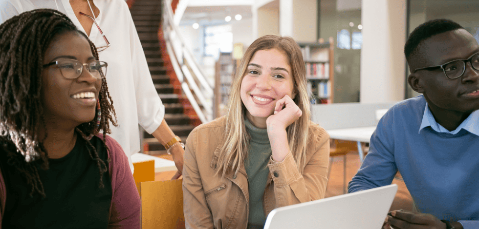 Trois étudiants assis autour d’une table dans une bibliothèque, souriants et engagés dans une discussion, avec un ordinateur portable ouvert devant eux – BSB