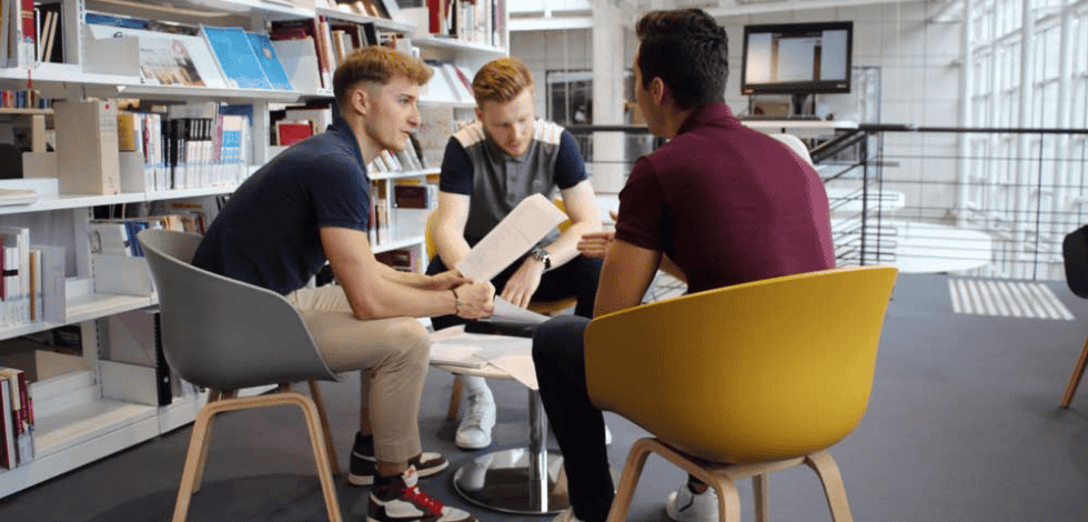 Trois étudiants assis dans une bibliothèque moderne, discutant et travaillant ensemble autour de documents posés sur une petite table – BSB