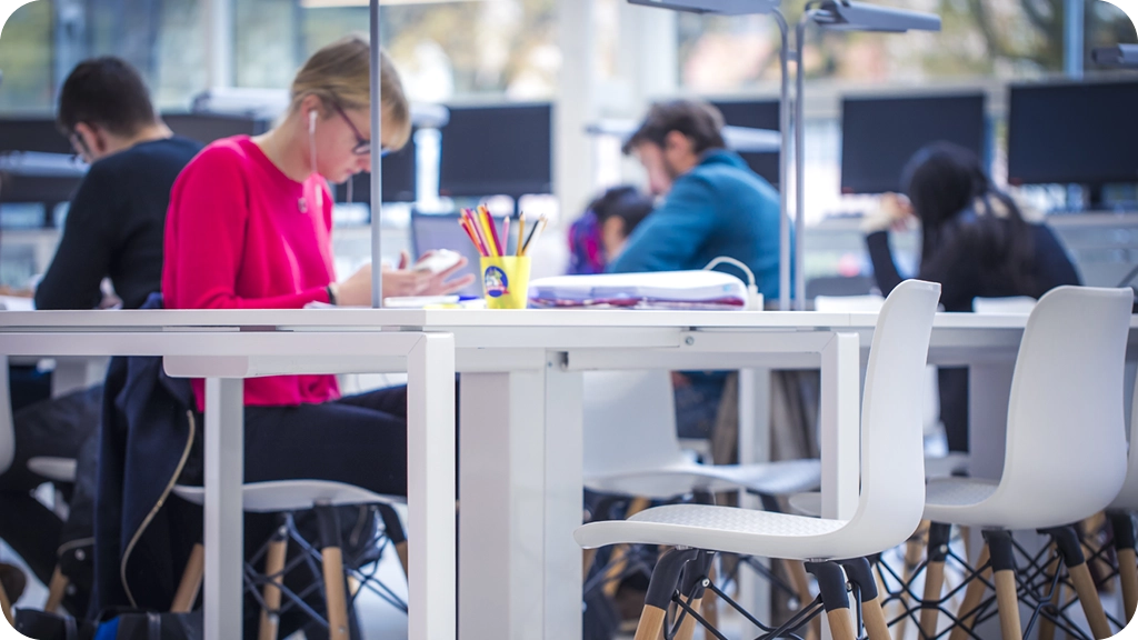 Des étudiants travaillent dans un espace de travail lumineux, assis à des tables blanches, certains concentrés sur leurs notes ou leurs écrans, avec du matériel scolaire posé sur les bureaux. – BSB 
