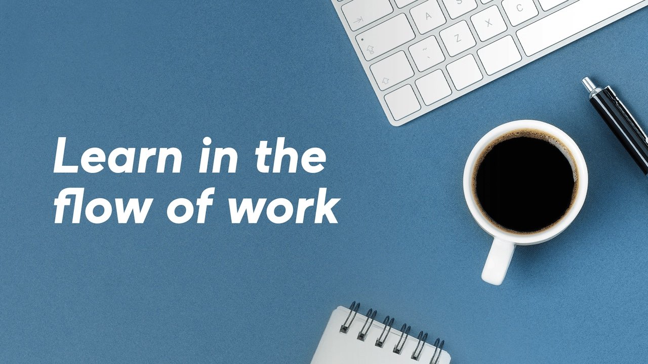 En blå bordplade, der kan ses oppefra, med et hjørne af et tastatur, en kaffekop og teksten "Learning in the flow of work" skrevet med store hvide bogstaver.