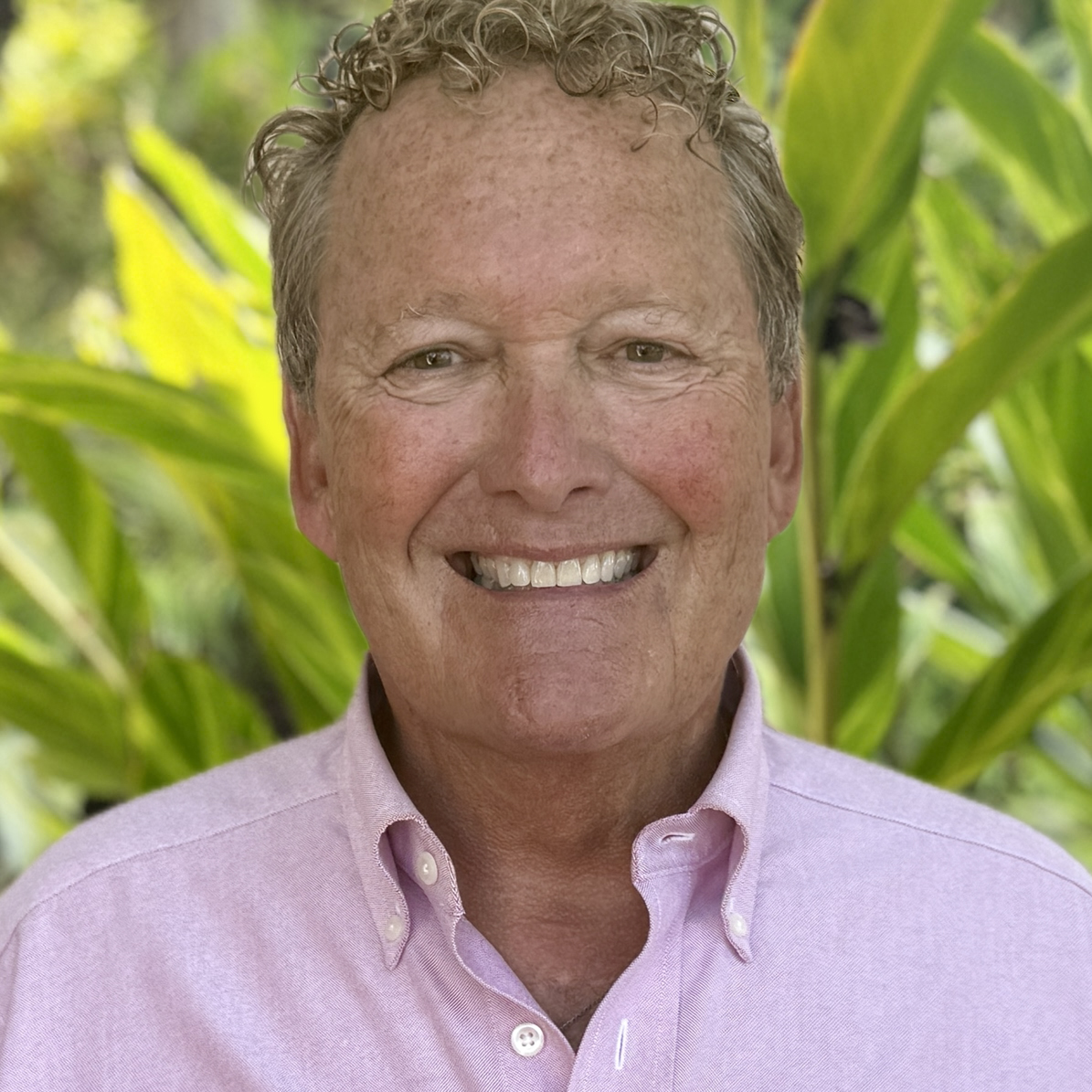 A man with grey curly hair smiling with a light blue shirt.