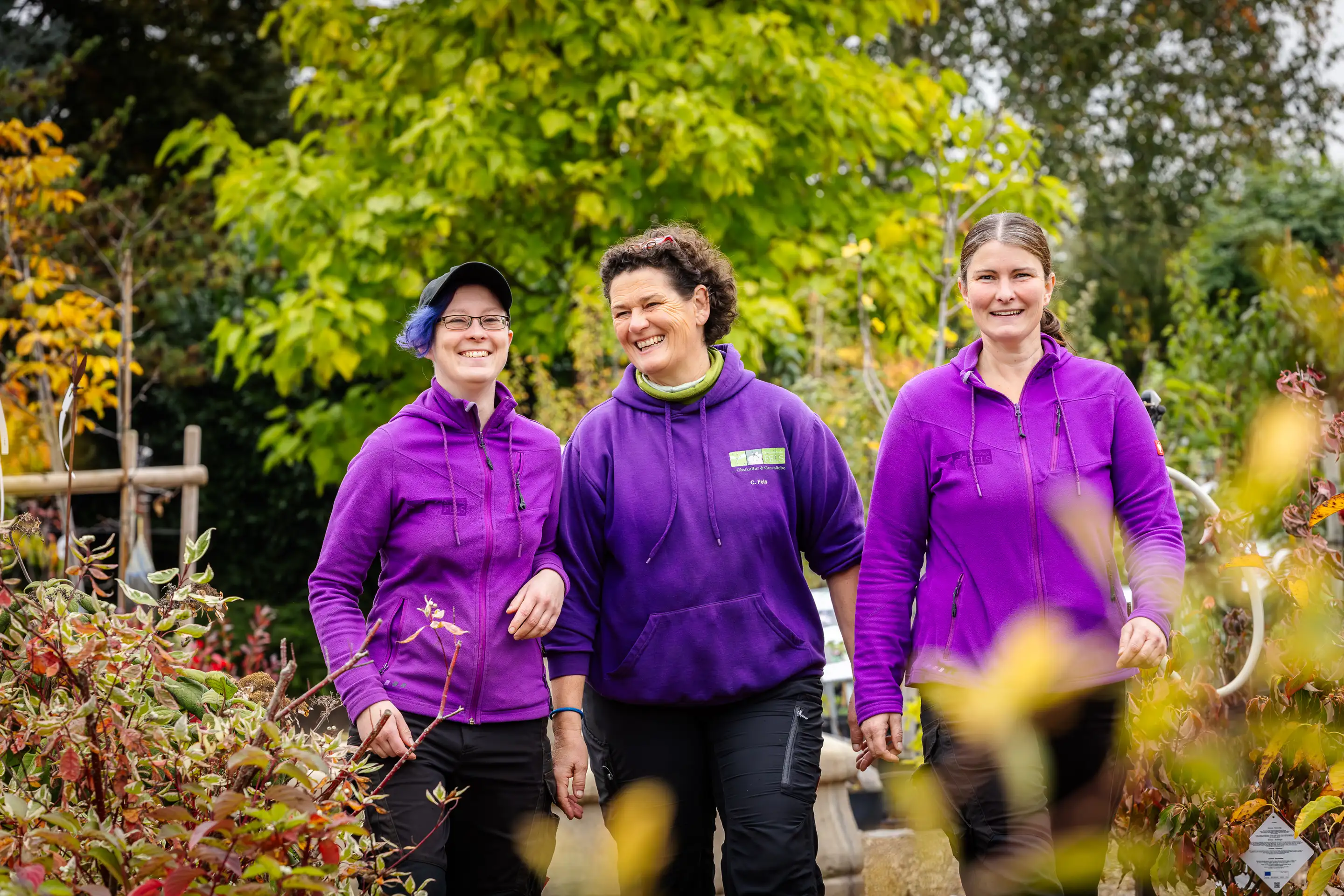 Drei Frauen in lila Jacken lachen und gehen in einer grünen Gartenumgebung spazieren.