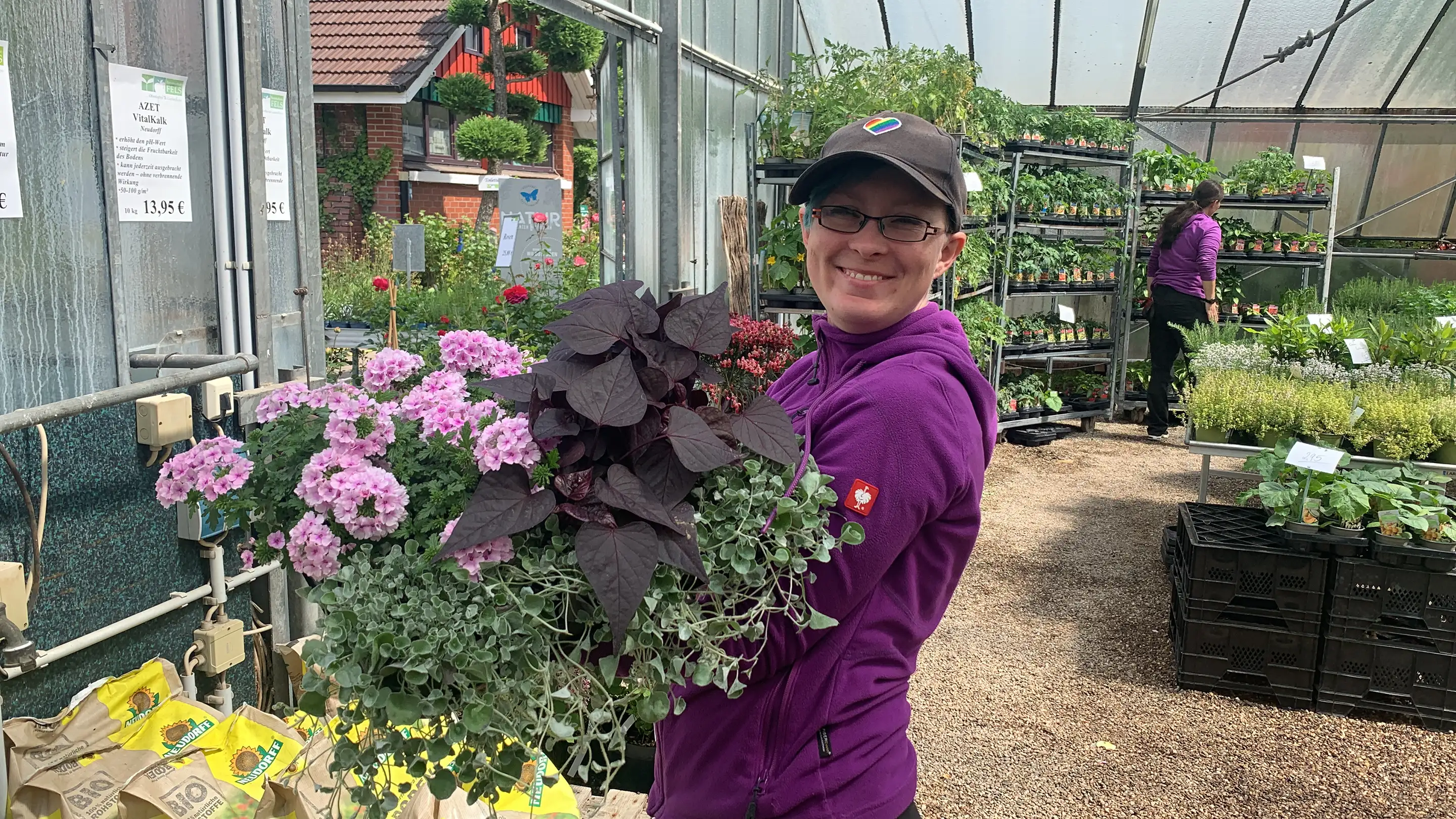 Lächelnde Person in violetter Jacke hält im Gewächshaus einen großen Blumenstrauß mit rosa und dunkelvioletten Blättern.