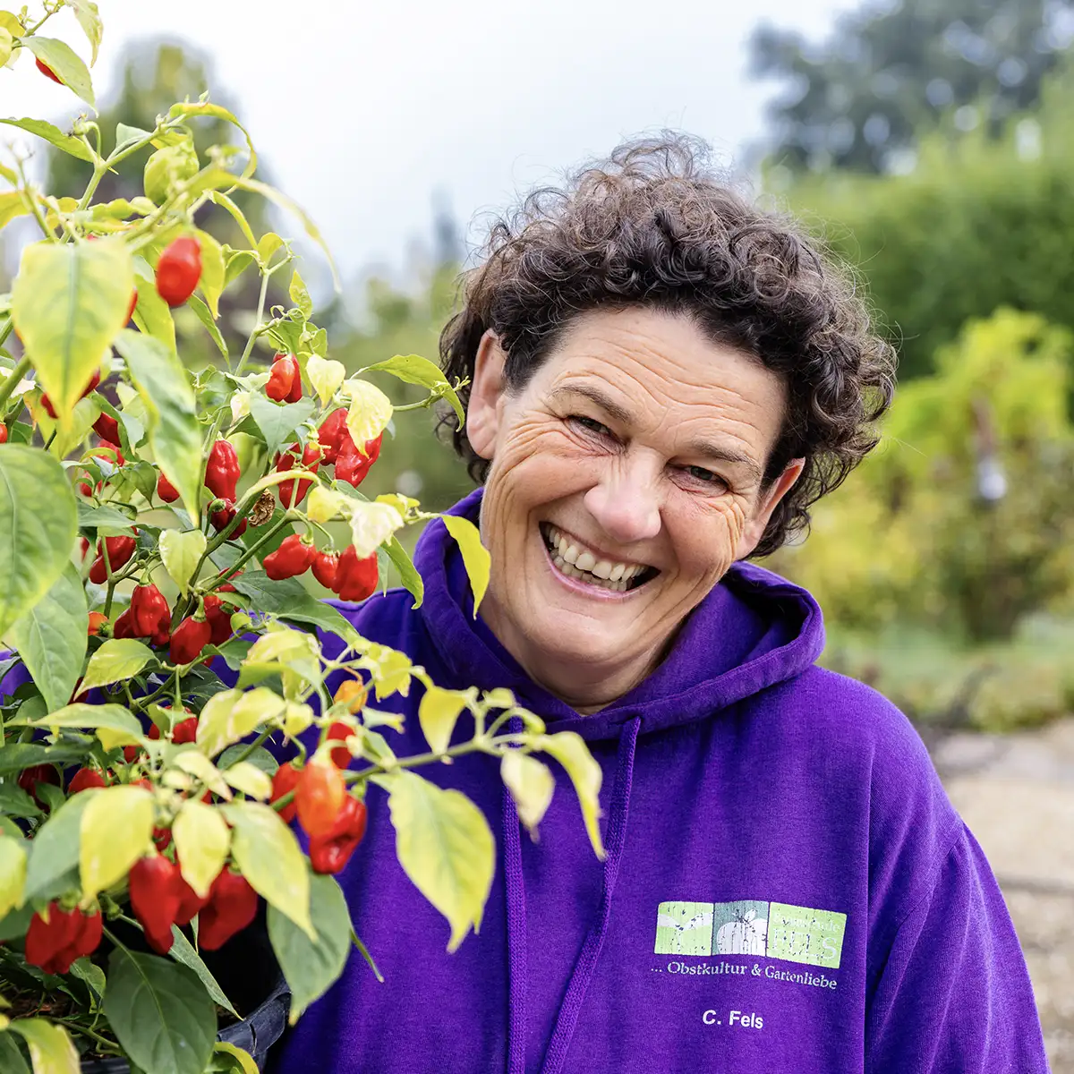 Frau mit lockigem Haar lacht neben Pflanze mit roten Chilischoten, trägt lila Kapuzenpullover.