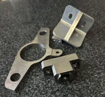This is a collection of many small parts that KT Machining has created. The part on the far left has a large hole with two small holes on either side of the large on, as well as a tiny hole below it. It is a T-shaped piece. The component in the top right is a small bracket with two holes and a support beam in the middle of the bracket. The component on the bottom right is shaped like a T with 3 holes, all on the bottom surfaces of the component.