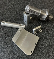 A collection of various metal component samples from KT Machining. The double-sided screw, a flat piece with a slight taper at the bottom, a cylindrical rod with holes, and two bracket type pieces are visible.